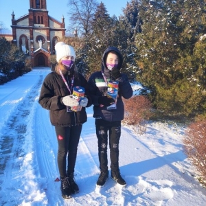 Dwoje wolontariuszy WOŚP stoi na zaśnieżonej drodze, w oddali widać budynek kościoła.