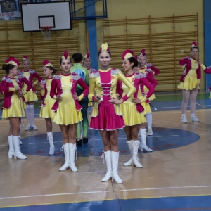 Grupa Mażoretek w strojach żółto-fioletowych stoi w sali gimnastycznej.
