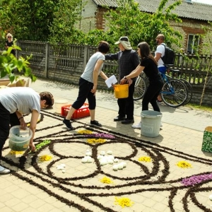 Trzy kobiety wysypują kwiaty na narysowanym wzorze. Obok stoi mężczyzna. W głębi widać zabudowania.