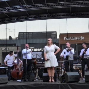Na scenie sześciu muzyków gra na instrumentach. Na przodzie kobieta w białej sukience trzyma w dłoni mikrofon.