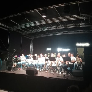 Na scenie orkiestra siedzi na krzesłach i gra. Wszyscy ubrani w białe bluzki.