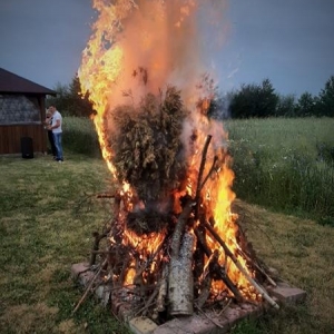 Na pierwszym planie palące się duże ognisko. W głębi widać mężczyznę, który stoi przy drewnianej altanie. 