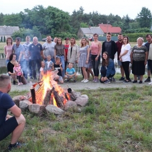 Po lewej stronie mężczyzna zwrócony plecami siedzi przy ognisku, w głębi grupa osób pozuje do zdjęcia. Ognisko otoczone jest kamieniami