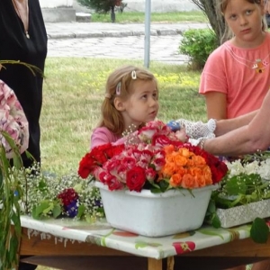 Dwie dziewczynki - mała i duża słuchają mówiącej kobiety.Dziewczynki stoją przy stole, na którym leżą kwiaty.