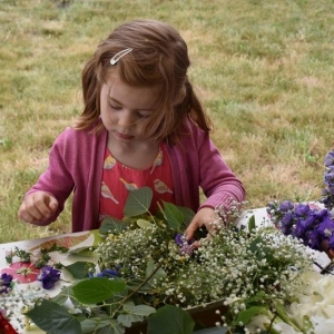 Mała dziewczynka stoi przy stole, na którym są kwiaty i układa z nich wianek.