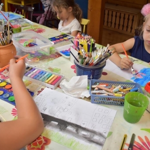 Na pierwszym planie dziewczynka maluje na kartonie.Na stole leżą farby, kredki,pędzelki.