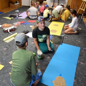 W dużej sali grupa dzieci siedzi na podłodze i wycina z kartonów. Na podłodze poukładane są różne kartony.W głębi widać sztalugi malarskie.