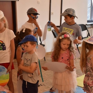 Grupa dzieci stoi przy siedzącej kobiecie .Dzieci trzymają w dłoniach papierowe kapelusze.