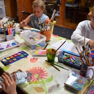Na stole leżą farby, kredki, pędzle.Dwie dziewczynki siedzą przy stole i  malują farbami na kartonie.