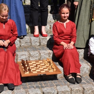 Grupa osób stoi na schodach i pozuje do zdjęcia. Dwie dziewczynki i małych chłopiec siedzą na schodach , obok nich leży plansza szachowa.W tle widać zamek z czerwonej cegły.Część osób ubranych jest w średniowieczne stroje.