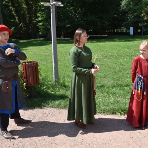 Na tle parkowych drzew stoją trzy osoby ubrane w średniowieczne stroje.