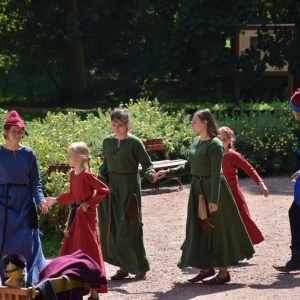 Grupa dzieci i młodzieży w średniowiecznych strojach tańczy na parkowym dziedzińcu.