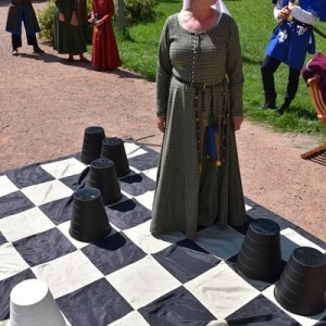 Na szachowej planszy stoi kobieta ubrana w średniowieczny strój.