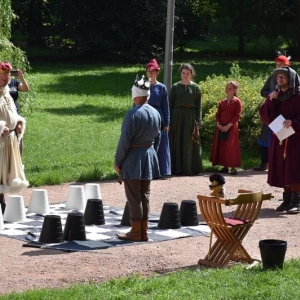 Dwóch mężczyzn w średniowiecznych strojach stoi na planszy szachowej, na której znajdują się rónież piony .Obok stoją osoby w średniowiecznych strojach, jeden z mężczyzn trzyma mikrofon i komentuje.