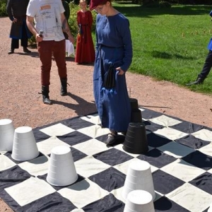 Na pierwszym planie na ziemi lezy plansza szachowa, na niej rozstawione sa piony.Na planszy stoi dziewczyna ubrana w strój średniowieczny, obok mężczyzna z mikrofonem.