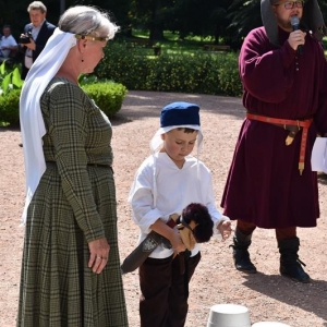 Obok kobiety w średniowiecznym stroju stoi mały chłopczyk, trzyma drewnianego konika.Na drugim planie mężczyzna z mikrofonem w dłoni.