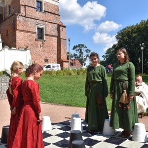 Na pierwszym planie plansza szachowa leżąca na ziemi, na planszy stoją cztery dziewczyny w strojach średniowiecznych.Obok siedzi mężczyzna w białym stroju.W tle mury zamku z czerwonej cegły.