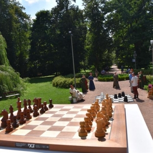 Na stoliku leży plansza szachowa z pionami, na dalszym planie widać dziedziniec parkowy, na którym tańczą osoby ubrane w średniowieczne stroje.