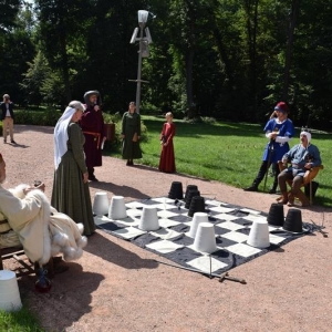 Plansza szachowa z pionami, po lewej i prawej stronie siedzą mężczyżni w strojach średniowiecznych.Na planszy stoi kobieta w stroju sredniowiecznym.