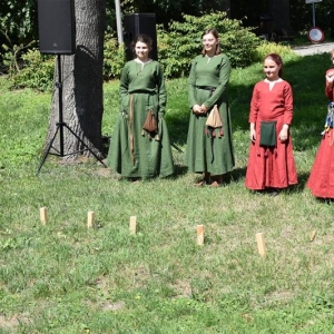 Cztery dziewczynki w średniowiecznych strojach stoją przy dużym drzewie, w dali drzewa parkowe.