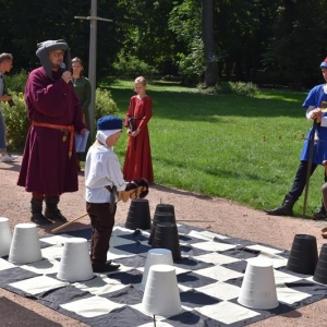 Na planszy szachowej znajduje się  mały chłopiec z drewnianym konikiem oraz duże piony szachowe.Obok mężczyźni i kobiety w strojach średniowiecznych, jeden z mężczyzn trzyma mikrofon i komentuje.
