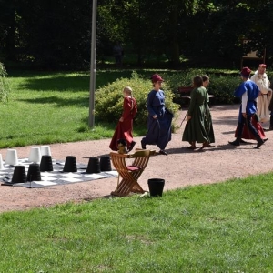 Grupa dzieci w średniowiecznych strojach tańczy na tle parku.Obok leży na ziemi plansza szachowa z rozstawionymi pionami.