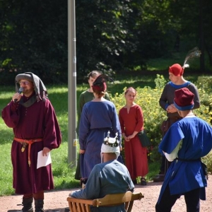 Grupa osób w średniowiecznych strojach stoi na tle drzew parkowych, jeden z mężczyzn mówi do mikrofonu, który trzyma w dłoni.