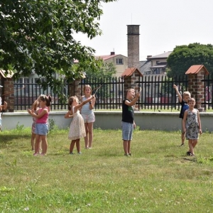 Grupa dzieci stoi na trawie , w dłoniach trzymaja patyki do których dowieszono wstążki, wstążki powiewają na wietrze.