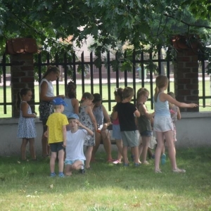 Grupa dzieci stoi na dworze przy wysokim drzewie.Każde dziecko przywiązuje do patyczka wstążkę.