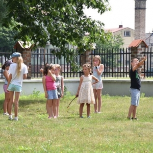 Grupa dzieci stoi na trawie i słucha osoby dorosłej.