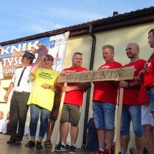 Na scenie stoi grupa kobiet i mężczyzn. Grupa w czerwonych koszulkach trzyma tablice drewnianą z napisem Wielenin.