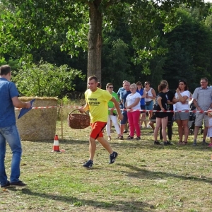 Piknik Sołtysów. Mężczyzna w żółtej koszulce biegnie z koszykiem. Po lewej stronie stoi mężczyzna, który mówi do mikrofonu. W tle drzewa i grupy ludzi