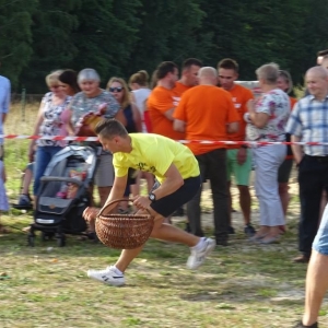 Piknik Sołtysów. Mężczyzna w żółtej koszulce biegnąc zbiera do koszyka ziemniaki.W tle stoi grupa ludzi.