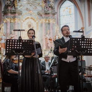 Koncert operowy w kościele. Kobieta i mężczyzna stoją przy mikrofonach i śpiewają.