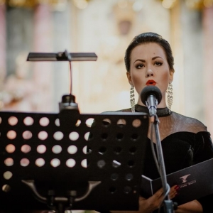 Kobieta ubrana w elegancką czarną suknie śpiewa do mikrofonu. W dłoniach trzyma nuty w czarnej obwolucie.