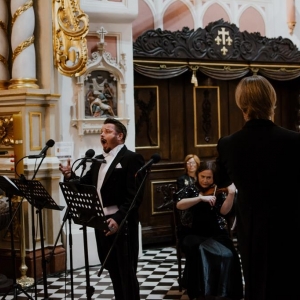 W kościele odbywa się koncert. Na pierwszym planie widać śpiewającego mężczyznę, obok siedzą muzycy. Po prawo stoi tyłem dyrygent.