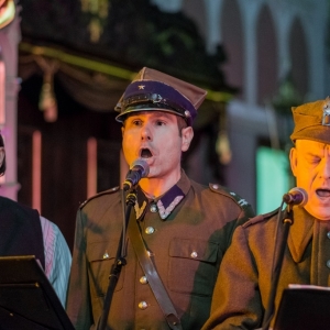 Trzej mężczyźni śpiewają do mikrofonu. Ubrani są w żołnierskie mundury.