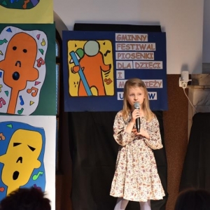 Dziewczynka w kolorowej sukience śpiewa na scenie. Za nią napis Gminny Festiwal Piosenki dla dzieci i młodzieży.