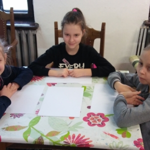 Uśmiechnięte trzy dziewczynki siedzą przy kolorowym stoliku, na stoliku białe kartony.