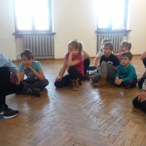 Grupa dzieci siedzi w okregu, słuchają mężczyzny w niebieskiej koszuli.