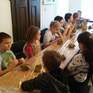 Grupa dzieci siedzi przy długim stole i lepi z gliny wazoniki.