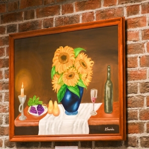Obraz w brązowej ramie zawieszony na ścianie z cegieł. Na obrazie narysowane są słoneczniki w niebieskim wazonie, owoce, zapalona świeca, kieliszek i butelka wina