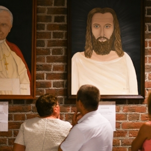Dwie kobiety i mężczyzna oglądają obrazy z Papieżem Janem Pawłem II i Panem Jezusem zawieszone na ścianie z cegieł