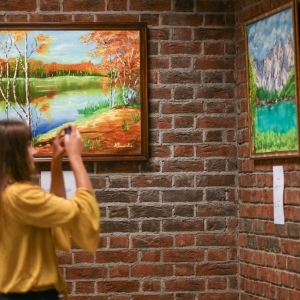 Kobieta w musztardowej bluzce robi zdjęcie obrazom zawieszonym na ścianie z cegieł. Na obrazach namalowane widoki z górami, drzewami i jeziorami