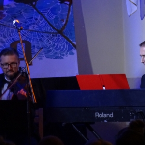 Dwóch mężczyzn gra na instumentach. Mężczyzna po lewej gra na skrzypcach, a mężczyzna po prawej gra na keyboardzie
