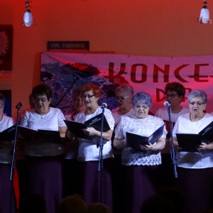 Grupa kobiet z zespołu śpiewaczego stoi na scenie i  śpiewa piosenkę