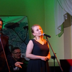 Kobieta i mężczyzna śpiewają na scenie. Na drugi planie dwóch mężczyzn gra na instrumentach