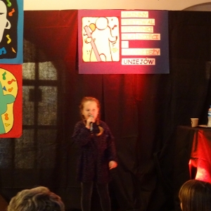 Dziewczynka w ciemnej sukience stoi na scenie i śpiewa piosenke
