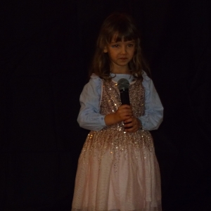 Dziewczynka w jasnej sukience stoi na scenie i trzyma mikrofon
