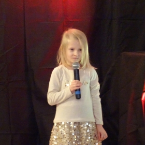 Dziewczynka w jasnej sukience z cekinowym dołem stoi na scenie i trzyma mikrofon